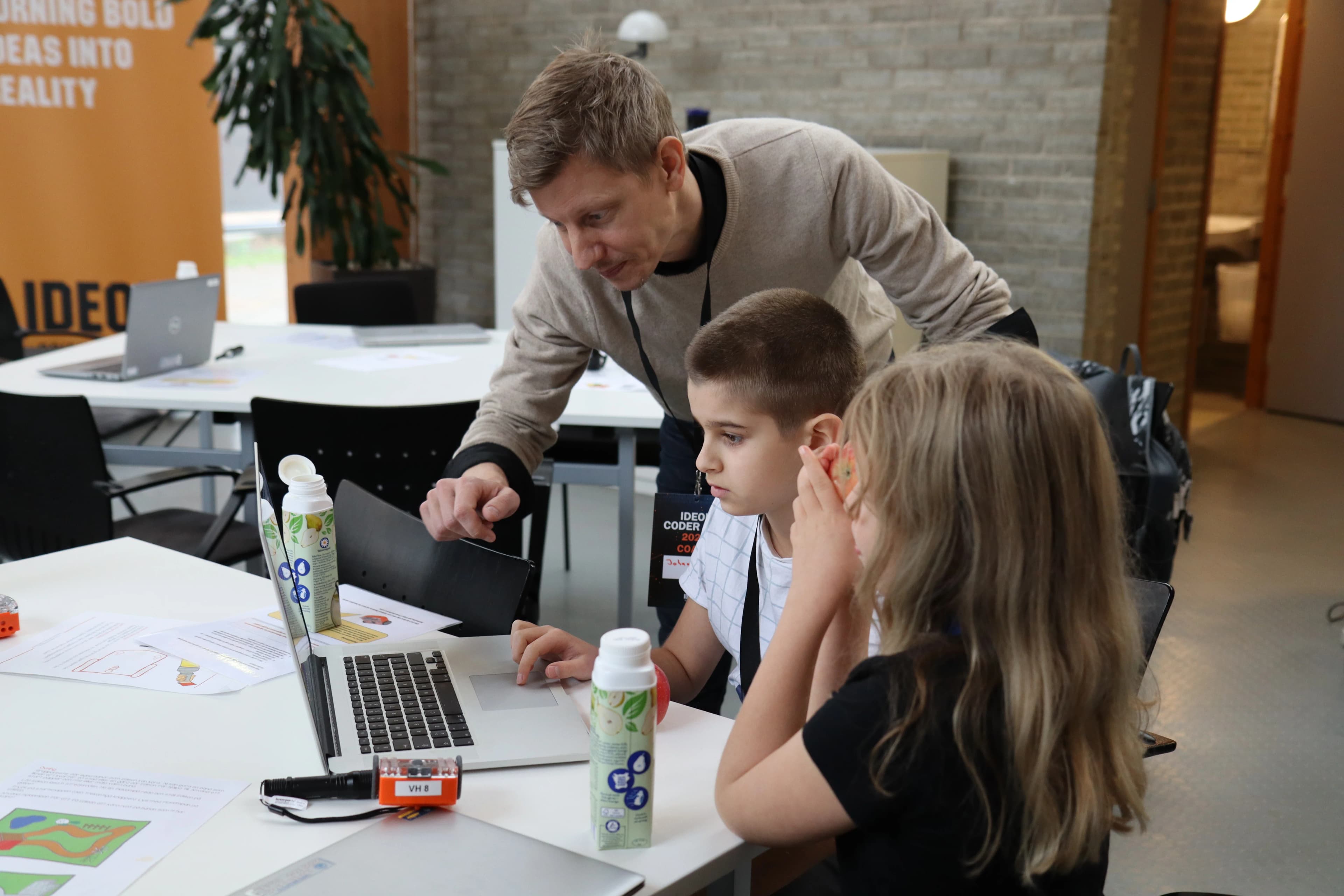 En coach lutar sig fram och hjälper två deltagare vid en laptop