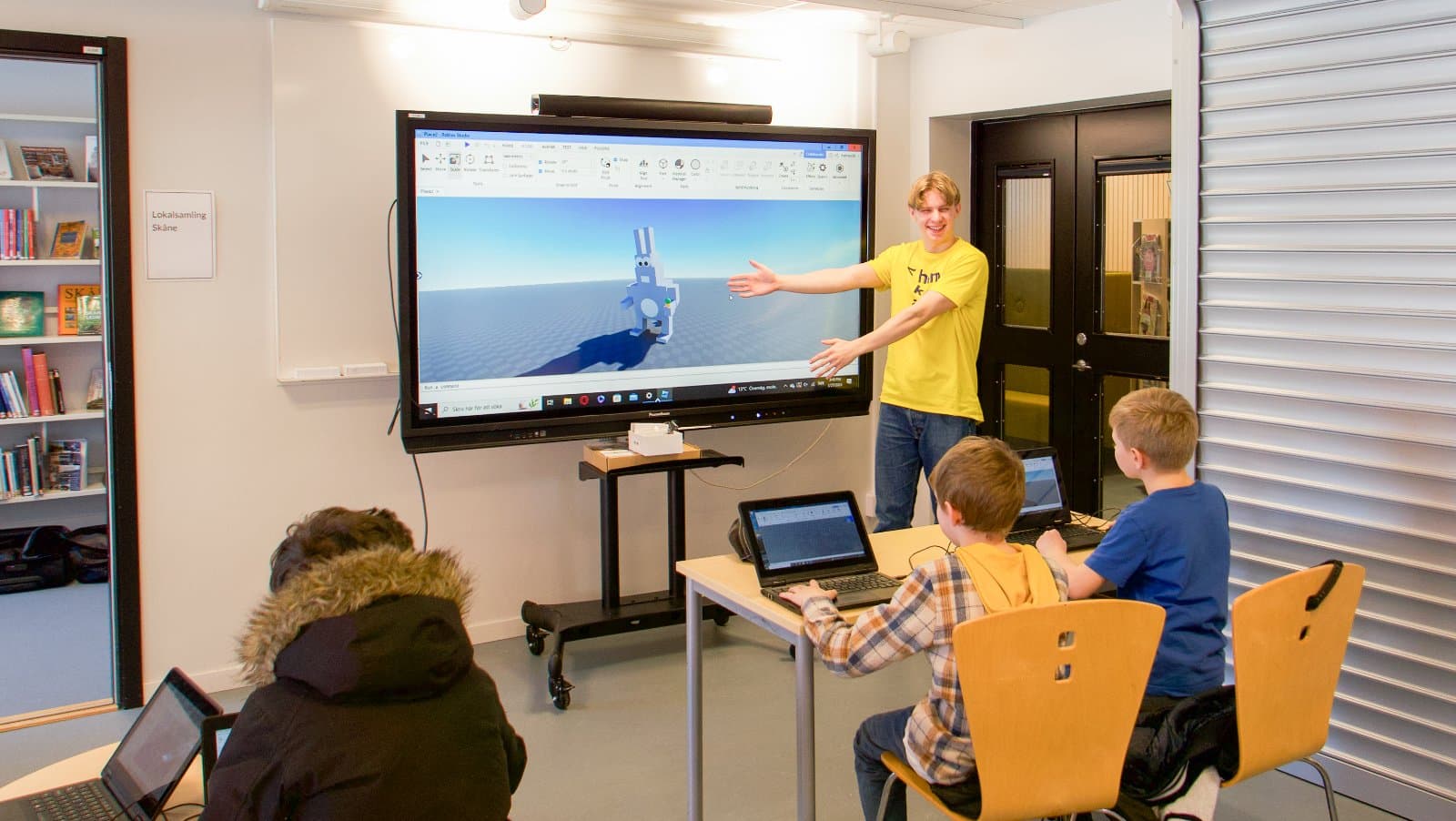 En ledare presenterar ett Roblox-bygge på storbildsskärmen medan barnen följer med vid sina datorer