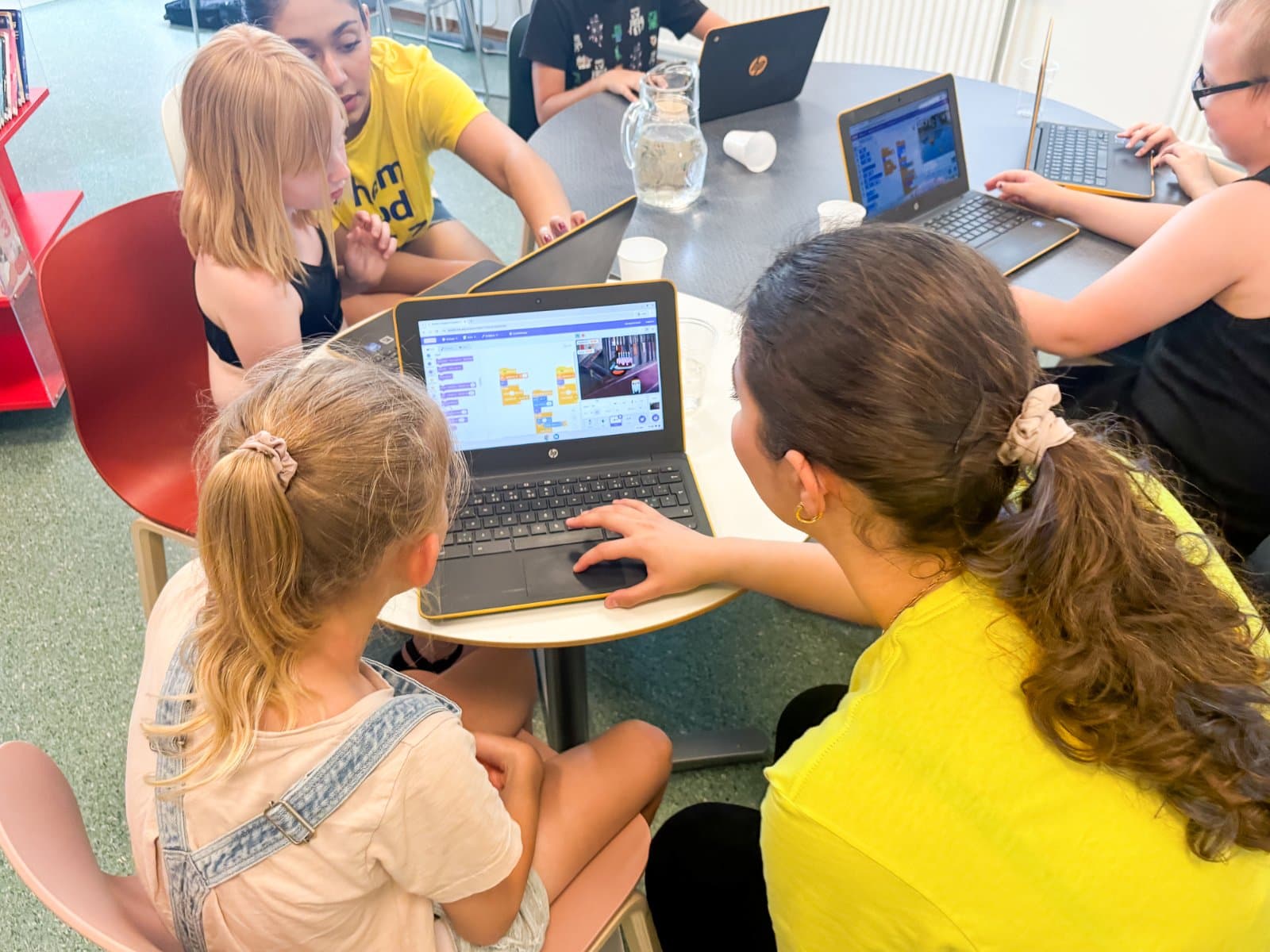 Barn programmerar i Scratch runt ett bord medan en ledare hjälper till