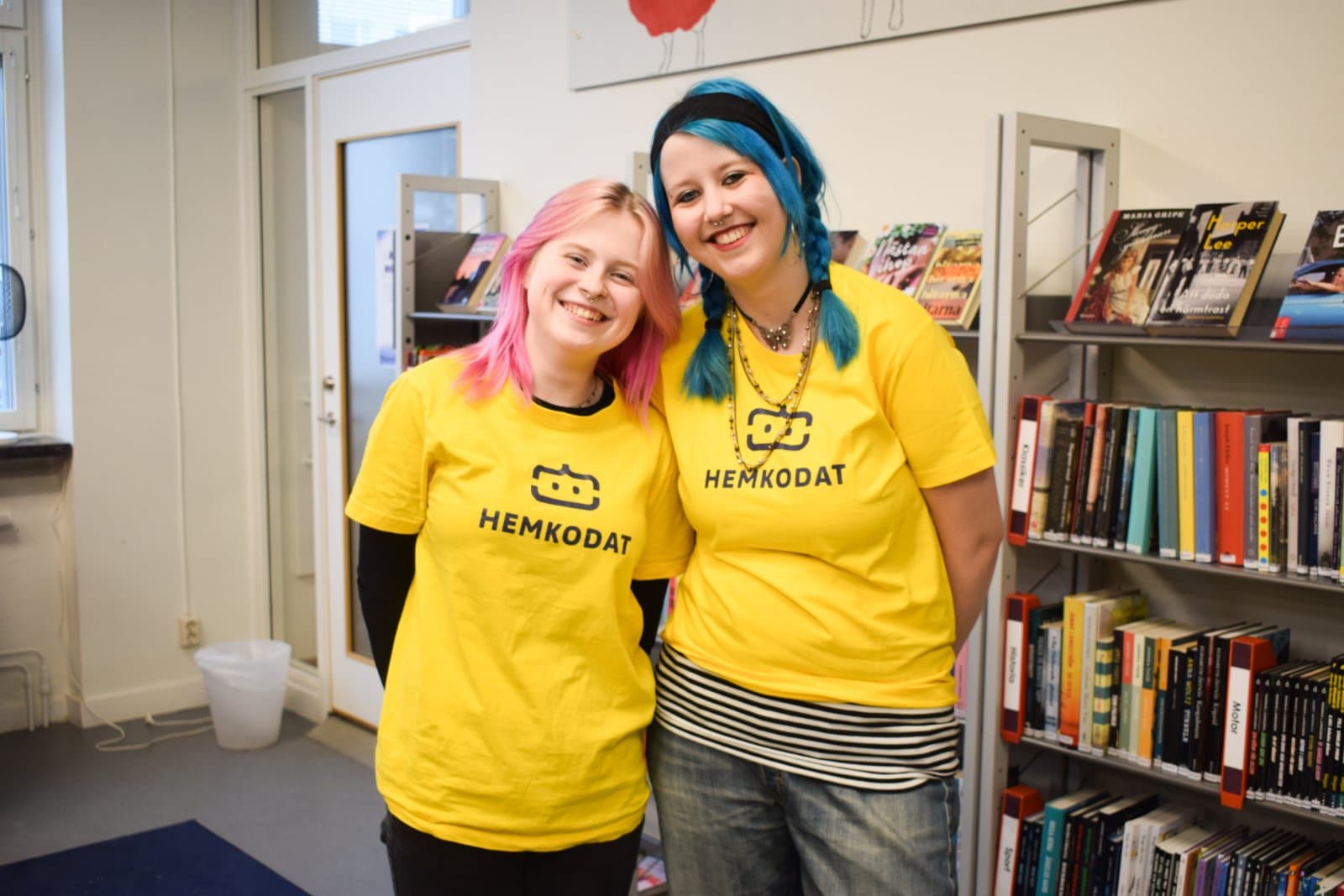 Två unga ledare i gula Hemkodat-tröjor står tillsammans i ett bibliotek